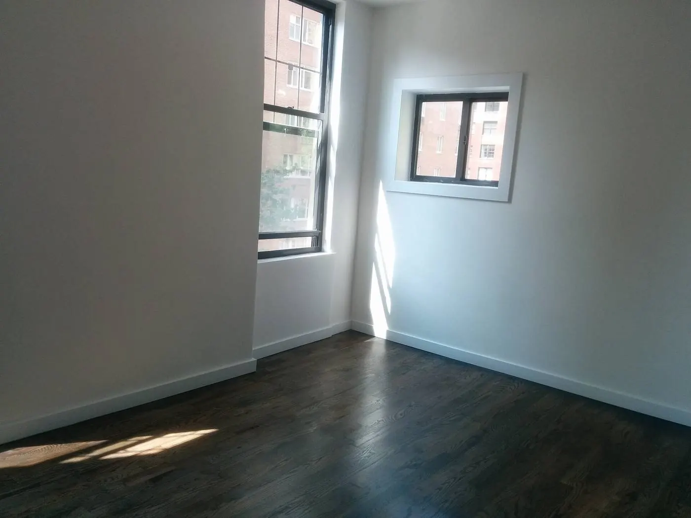 Empty sunlit room with two corner windows and a dark wooden floor