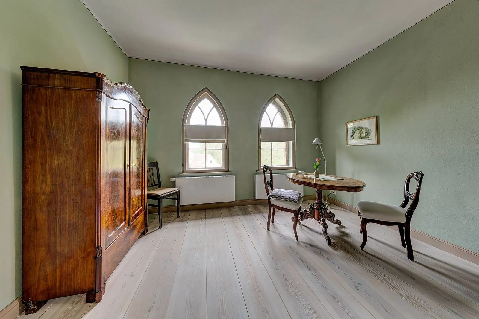 Bright room in an old house: soft green walls, pointed-arch windows, wide floorboards and an antique wardrobe